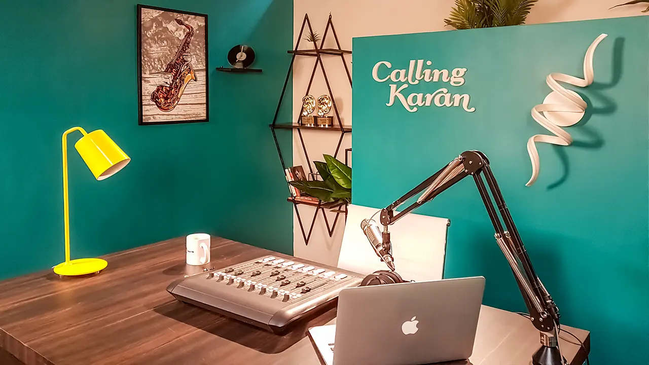 Calling Karan Season 2 radio booth setup with teal wall, console, mic arm, laptop, yellow lamp and award shelf