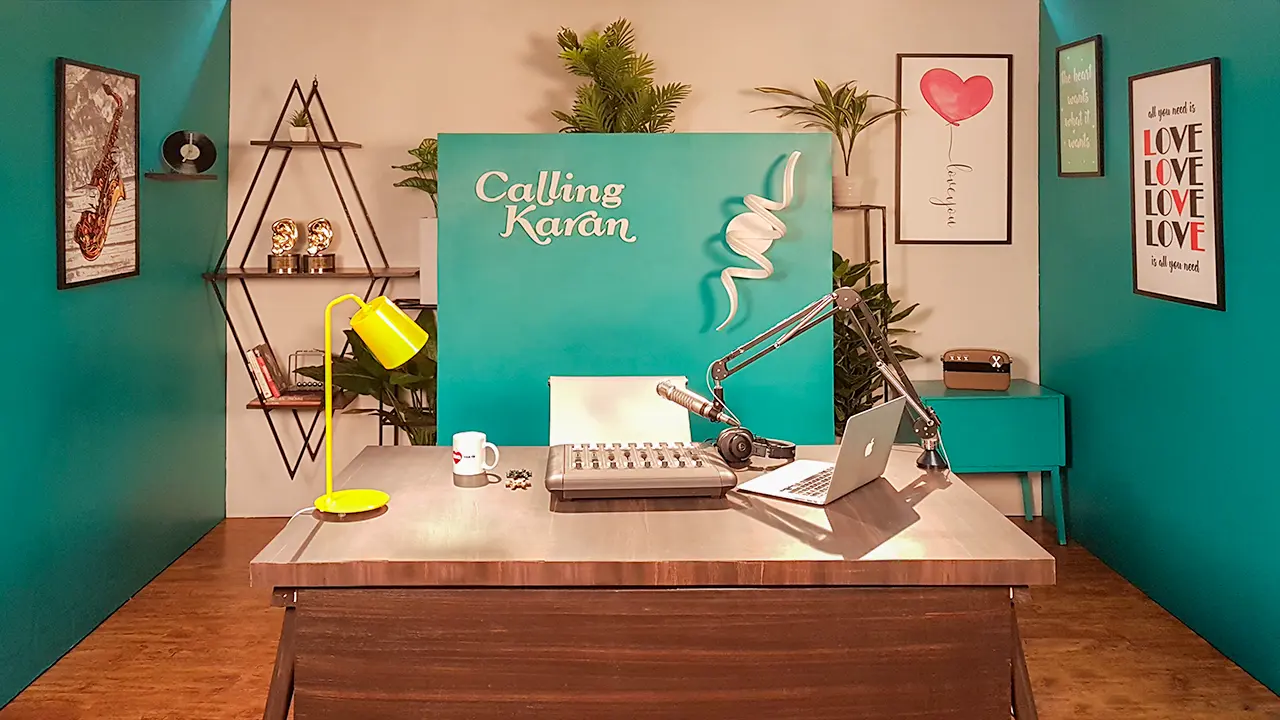 Calling Karan Season 2 radio show set with teal brand backdrop, desk mic, laptop, yellow lamp and bookshelf props