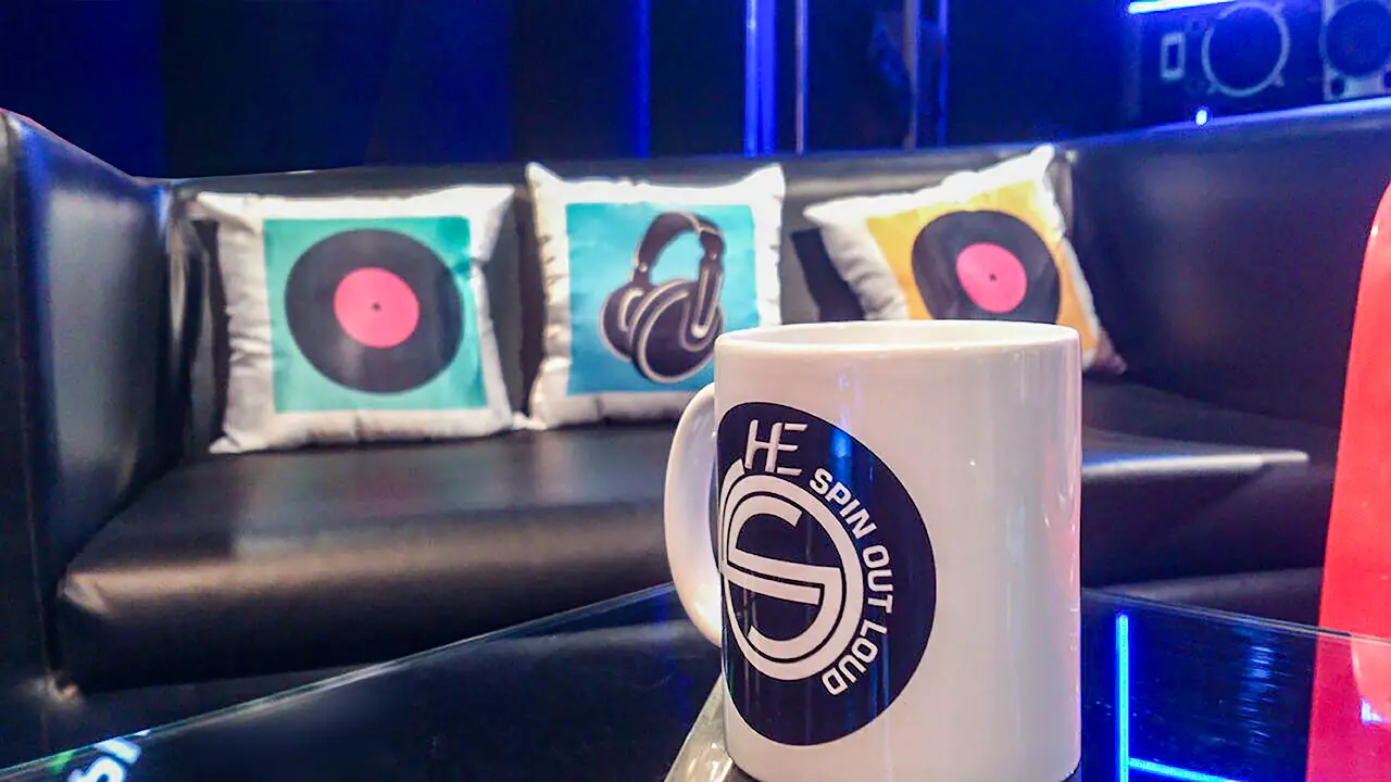 Branded HE Spin Out Loud coffee mug on lounge table with vinyl-themed cushions and leather seating in the background