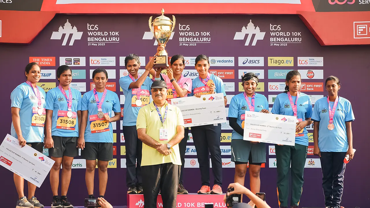 Winning women’s police teams on podium at TCS World 10K Bengaluru 2023, with medals, trophies, and cheques — sponsor logos and branding arranged as per guidelines, deliverables, and media visibility