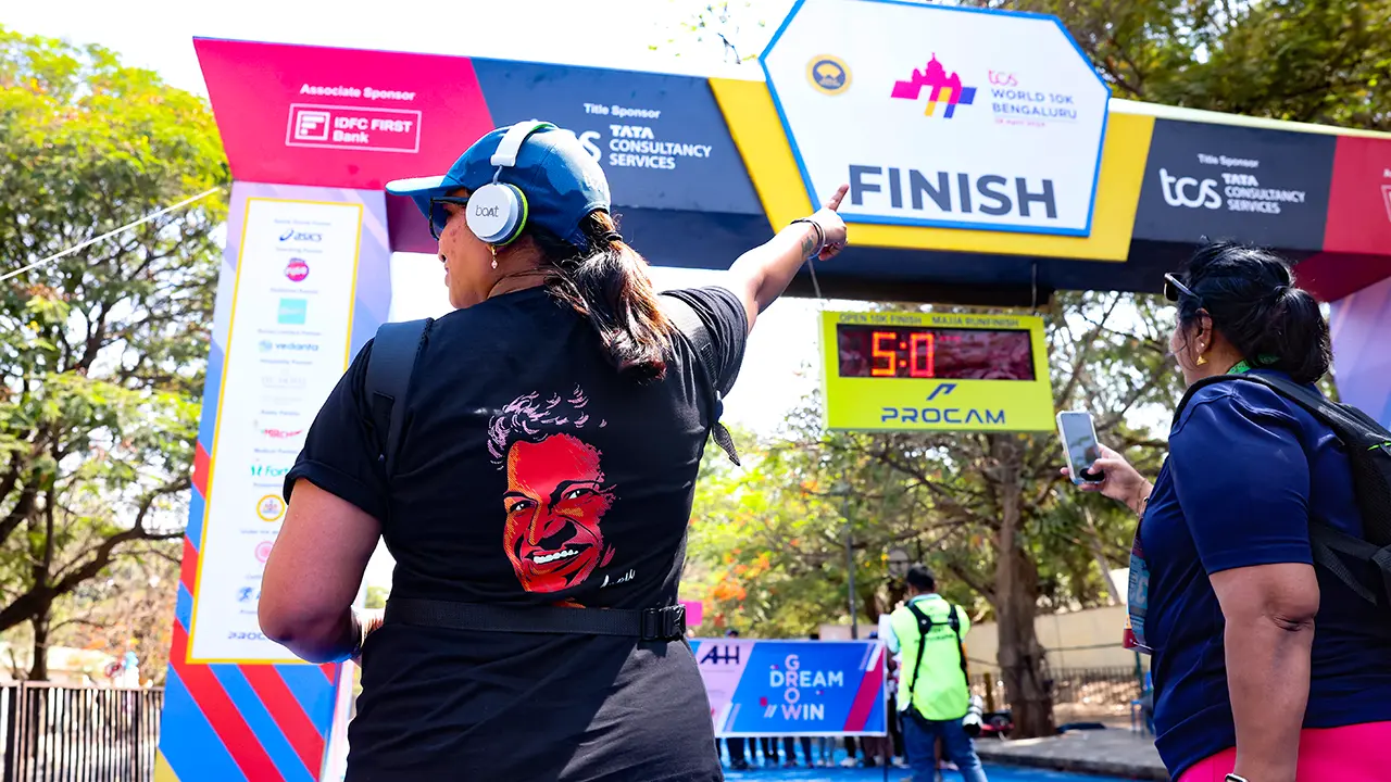 Participant celebrates at the TCS World 10K Bengaluru finish gate, pointing toward the Procam clock beneath bold sponsor branding and a Puneet Rajkumar mural on her t-shirt