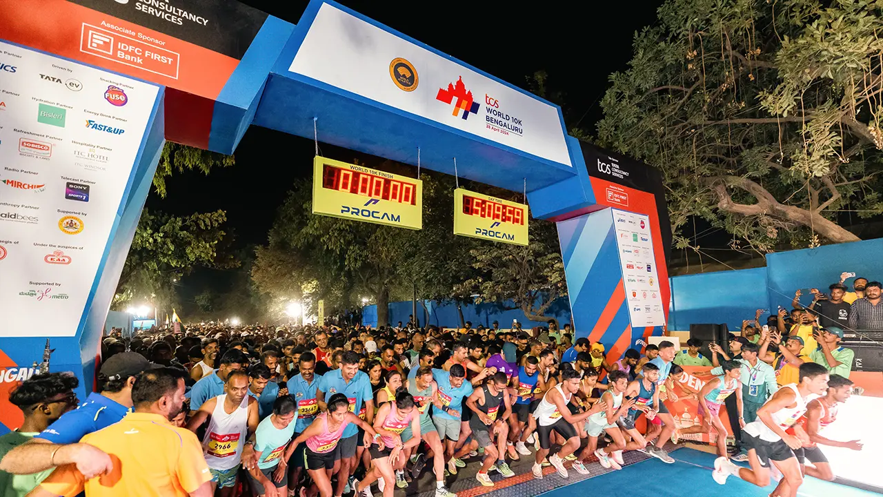 Mass start moment at TCS World 10K Bengaluru 2024, captured under a fully branded start gate featuring sponsor walls and twin digital timers